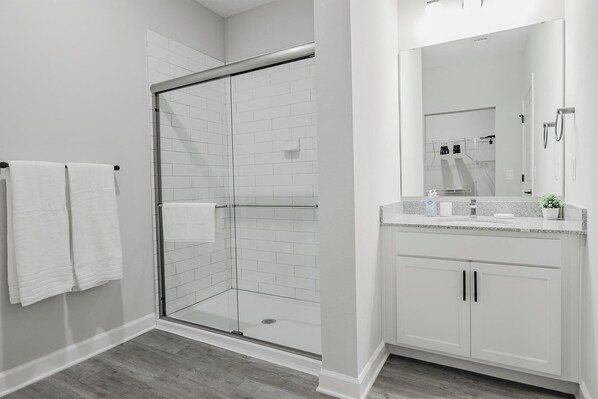A modern bathroom with a glass shower enclosure, white tiled walls, a granite countertop, and a large mirror. - Landing | Chic 2BD, Gym, Pool (Tice)