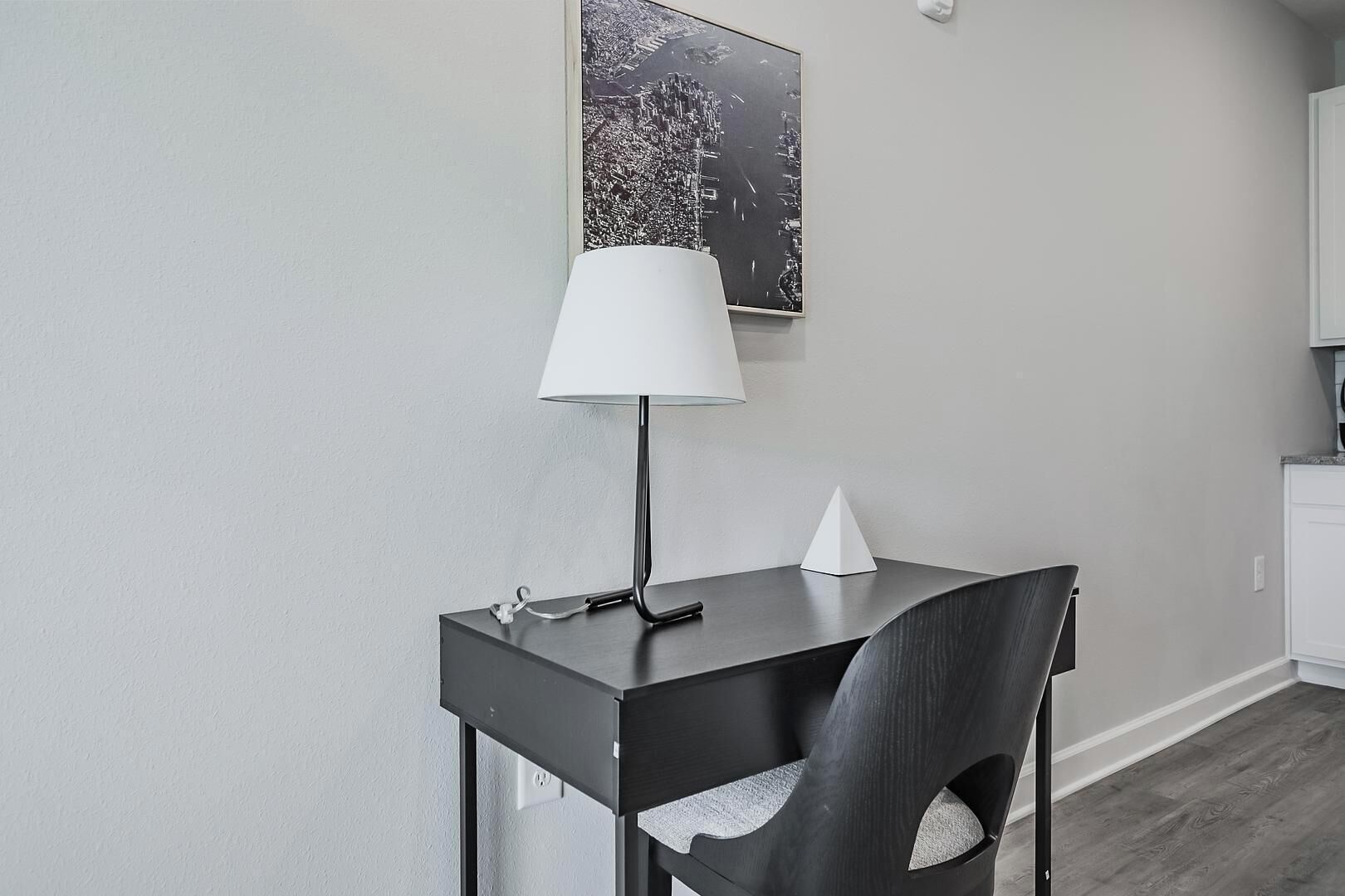 Une pièce moderne et minimaliste, avec un bureau en bois foncé, une lampe blanche et une chaise. Un tableau encadré est accroché au mur, et un meuble blanc se trouve au fond de la pièce.
