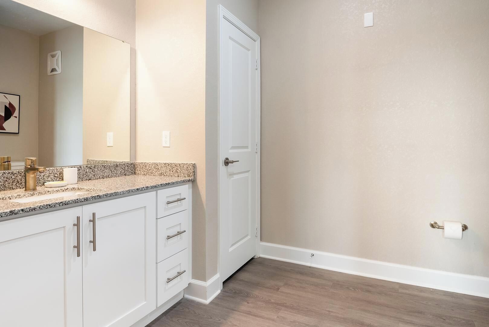 A bathroom with a granite countertop, white cabinets, a white door, and a wooden floor.