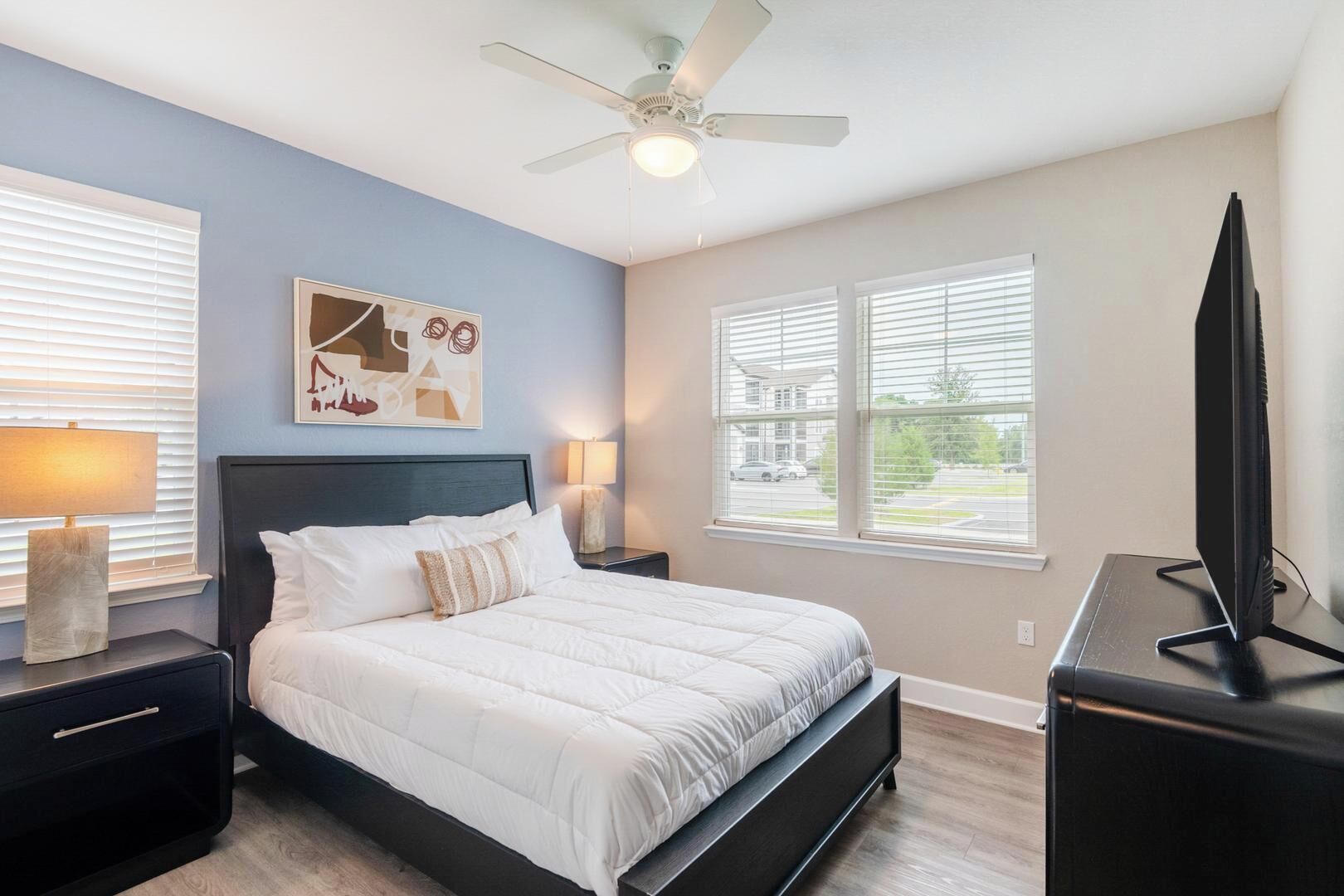 A bedroom with a bed, nightstands, a ceiling fan, a TV, and windows.