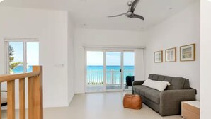 Un vaste salon doté d’un canapé gris, d’un pouf marron et d’un ventilateur de plafond. Une grande porte-fenêtre coulissante donne accès à un balcon offrant une vue sur l’océan.