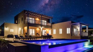 Une maison moderne à deux étages avec une piscine, illuminée la nuit sous un ciel étoilé.