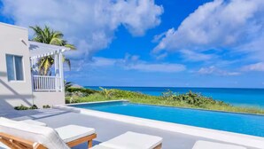 Un espace moderne aménagé au bord de la piscine, avec des chaises longues blanches et une vue dégagée sur l’océan.