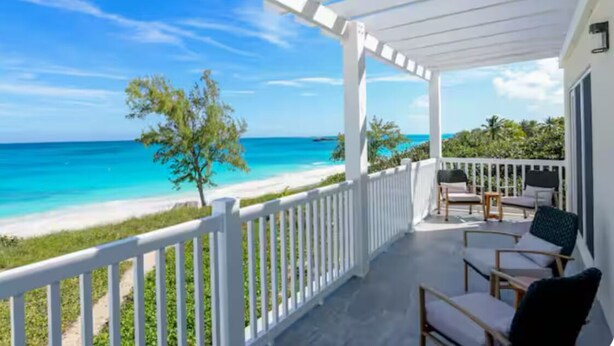 Un balcon abrité, doté de balustrades blanches, qui donne sur une plage aux eaux turquoise et limpides, avec un petit arbre.