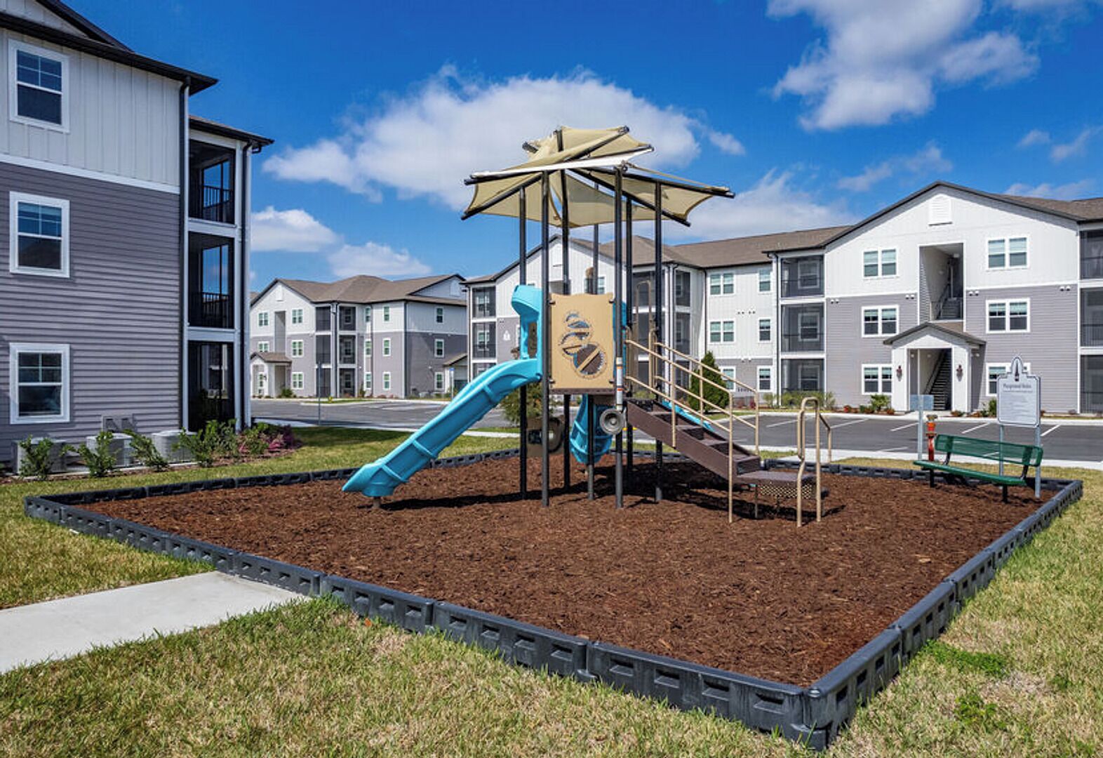 Ein Spielplatz mit Rutsche und Klettergerüsten, umgeben von einer Grünfläche mit Rindenmulch und Gras, vor dem Hintergrund von mehrstöckigen Wohnhäusern.
