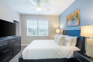 Une chambre d’hôtel avec un lit, une télévision, un ventilateur de plafond, une fenêtre avec des stores, un tableau au mur et une table de chevet avec une lampe.