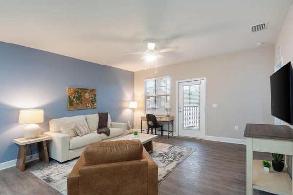 Un salon comprenant un canapé, un fauteuil marron, un bureau avec une chaise, un téléviseur et un ventilateur de plafond.