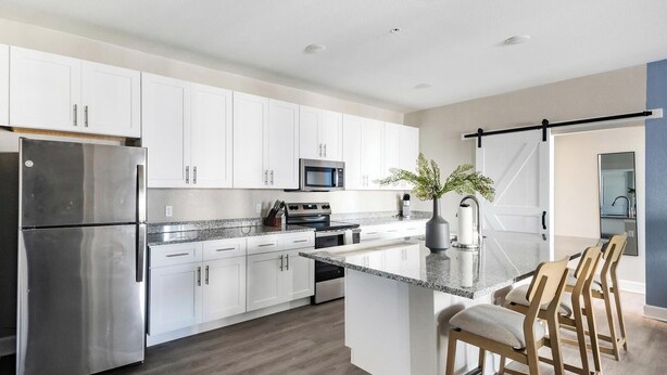 A modern kitchen with white cabinets, stainless steel appliances, a granite countertop, and a bar area with stools. - Landing | Beautiful 2BD, Gym, Pool (Ocala)