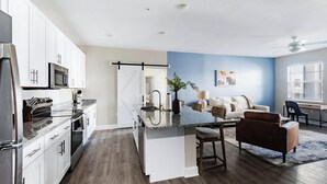 A modern kitchen with white cabinets, a granite countertop, and stainless steel appliances. There is a dining area with a bar stool and a sofa. - Landing | Beautiful 2BD, Gym, Pool (Ocala)