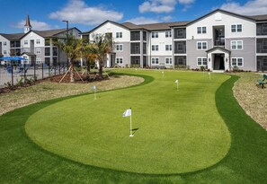 Un terrain de golf avec un drapeau sur le green, entouré d’immeubles à plusieurs étages, avec une aire de jeux et le clocher d’une église en arrière-plan.