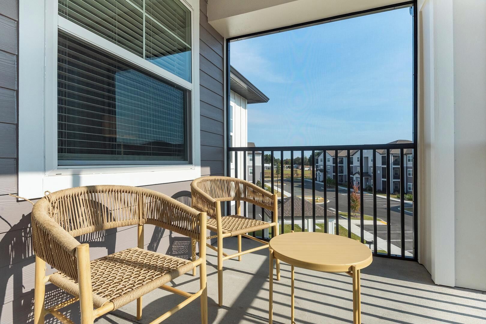 Un balcon avec des fauteuils en osier et une petite table ronde, donnant sur un parking et des immeubles.