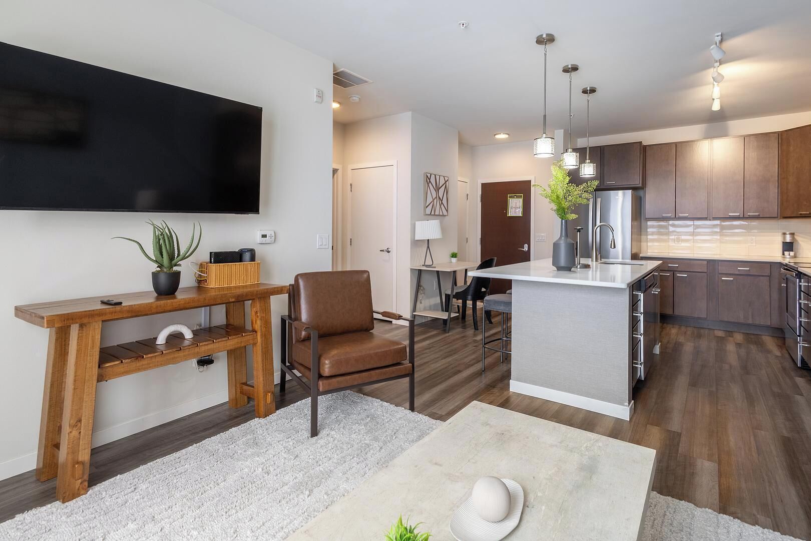 A modern kitchen with wooden cabinets, a central island, pendant lighting, and a flat-screen TV mounted on the wall.