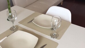 Ein Esstisch mit Geschirr, Besteck und Weingläsern, eine Vase mit Pflanze sowie eine Couch im Hintergrund.