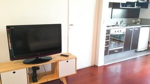 Ein Flachbildfernseher auf einem Holzständer mit Fernbedienung, eine weiße Tür und ein Küchenbereich mit Schränken und Geräten.