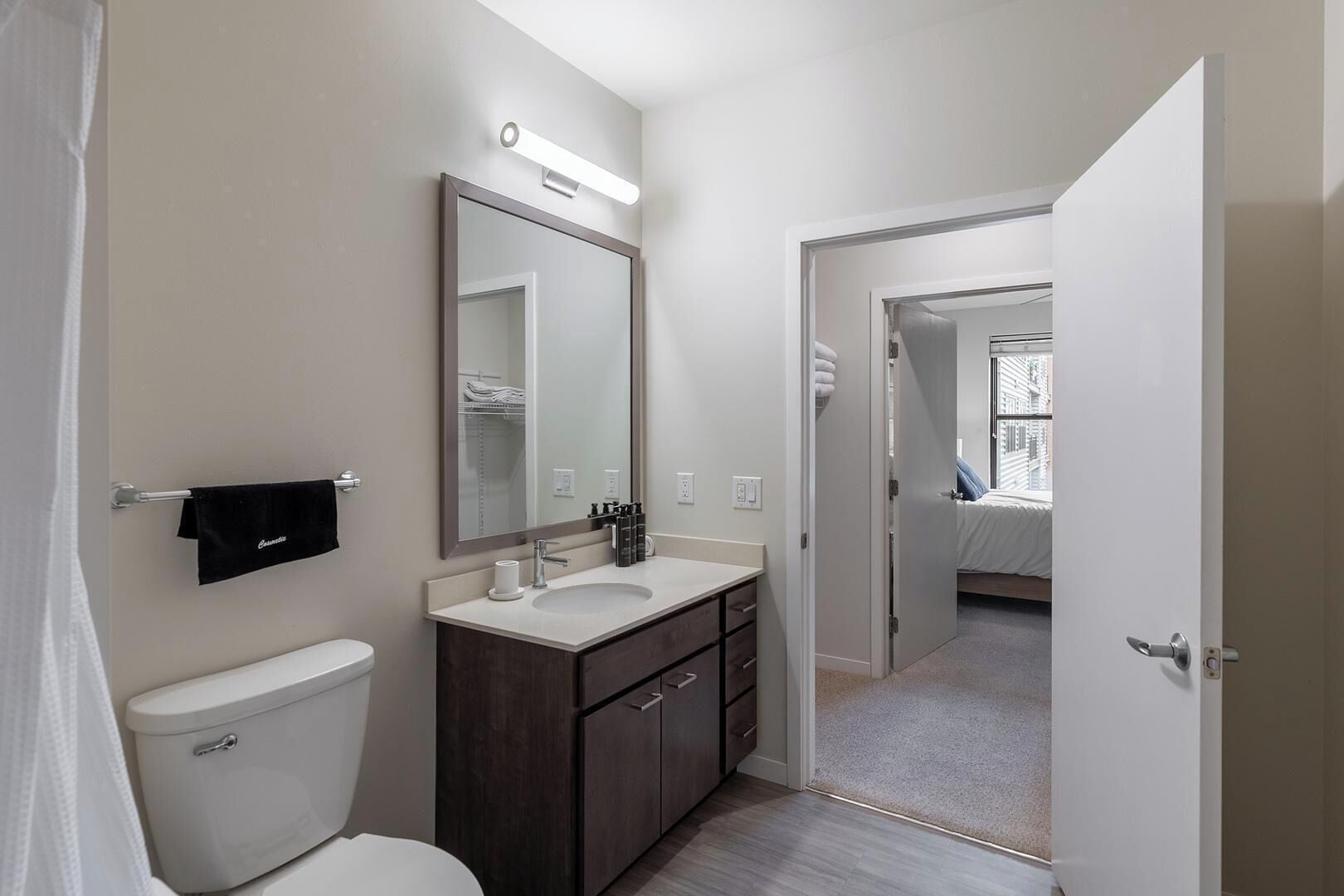 A modern bathroom with a toilet, sink, and mirror. There is a towel rack with a black towel and a mirror above the sink. The bathroom has a door leading to a bedroom with a bed and a window.