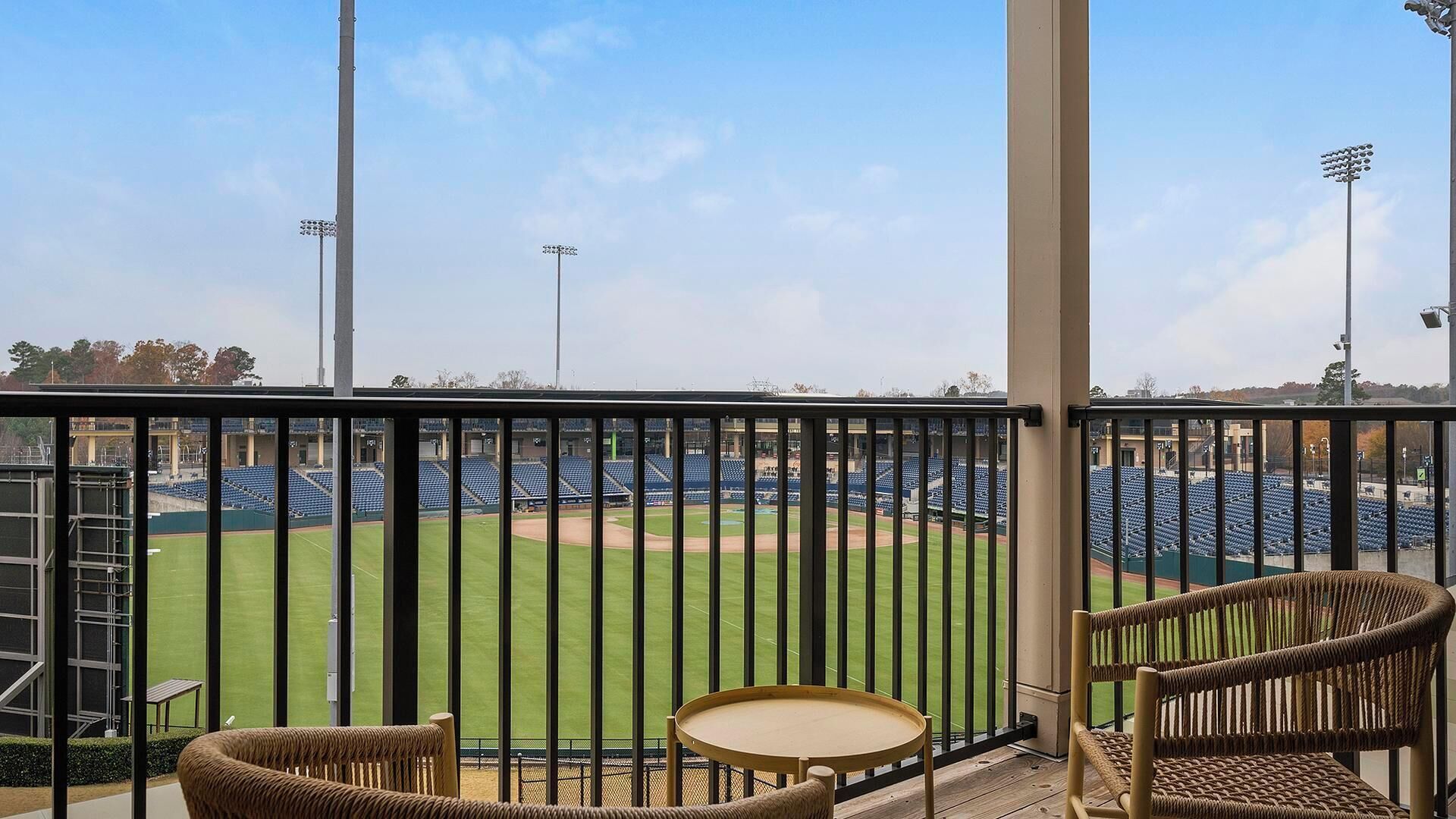 Un balcón con sillas de mimbre y una mesita pequeña, con vistas a un campo de béisbol con gradas vacías y focos.