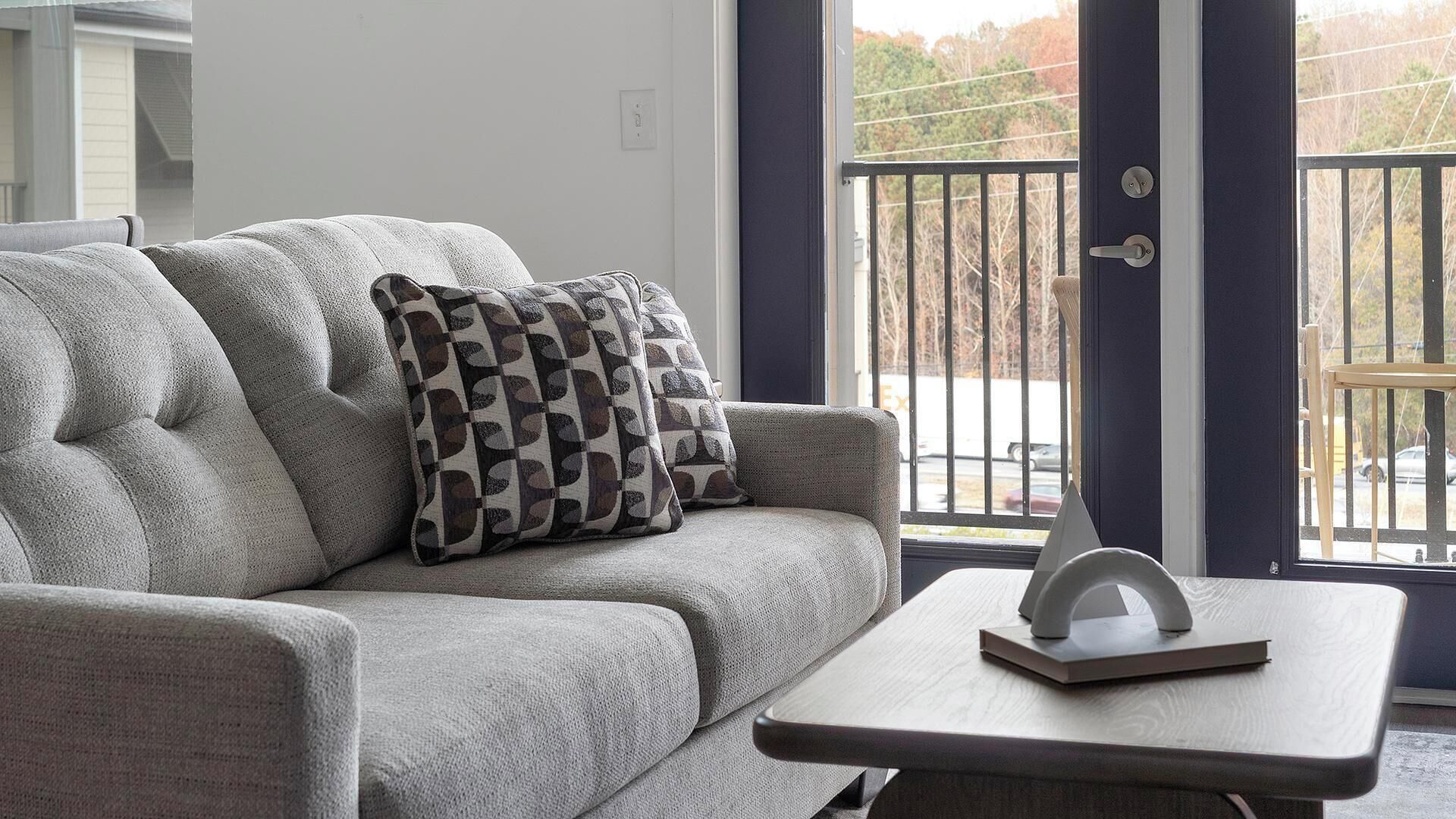 A grey sofa with a patterned pillow, a small wooden table with a book and a decorative object, and a view of a balcony with a railing.