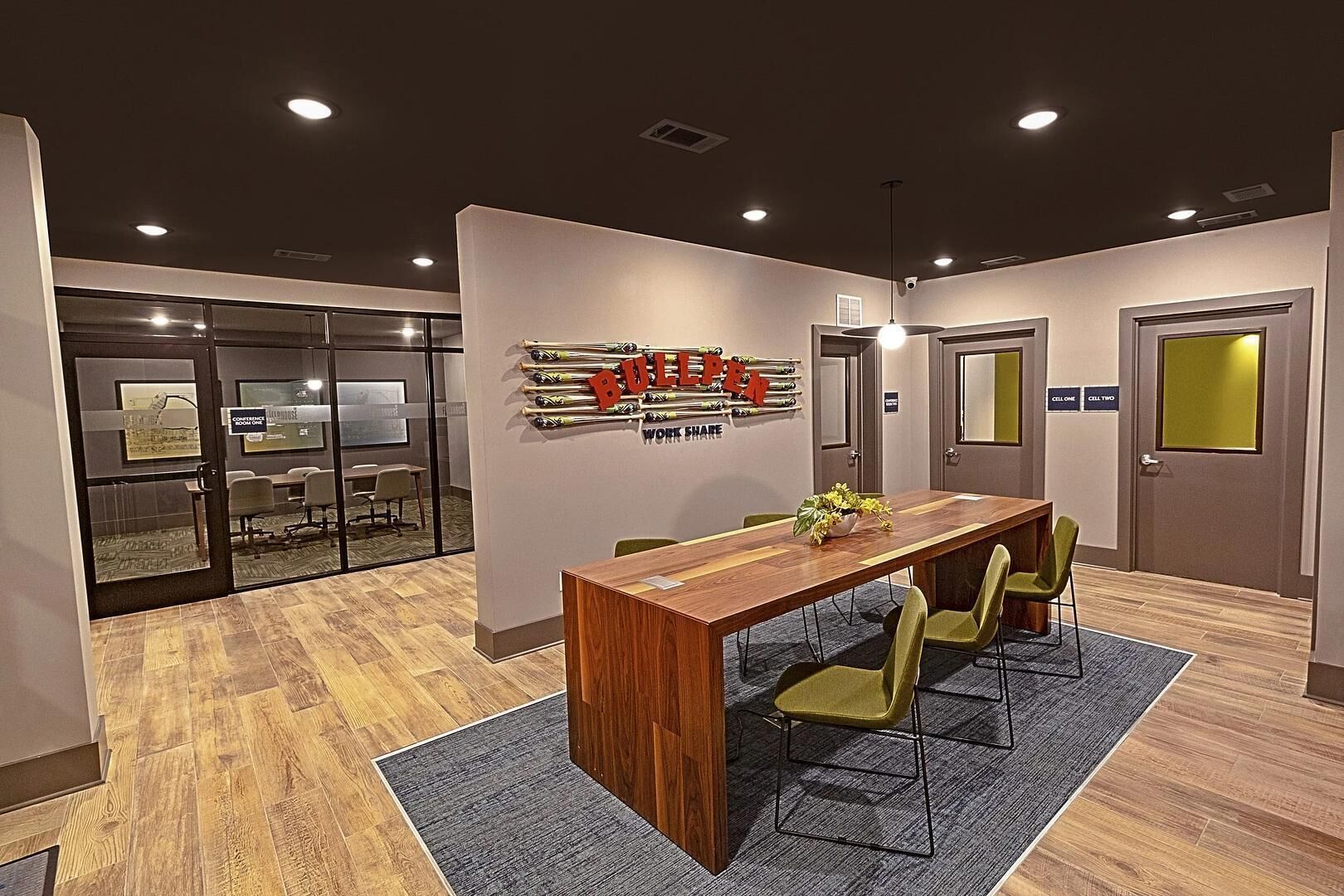 A conference room with a long wooden table, green chairs, a wall-mounted bat display, and a gray carpet.