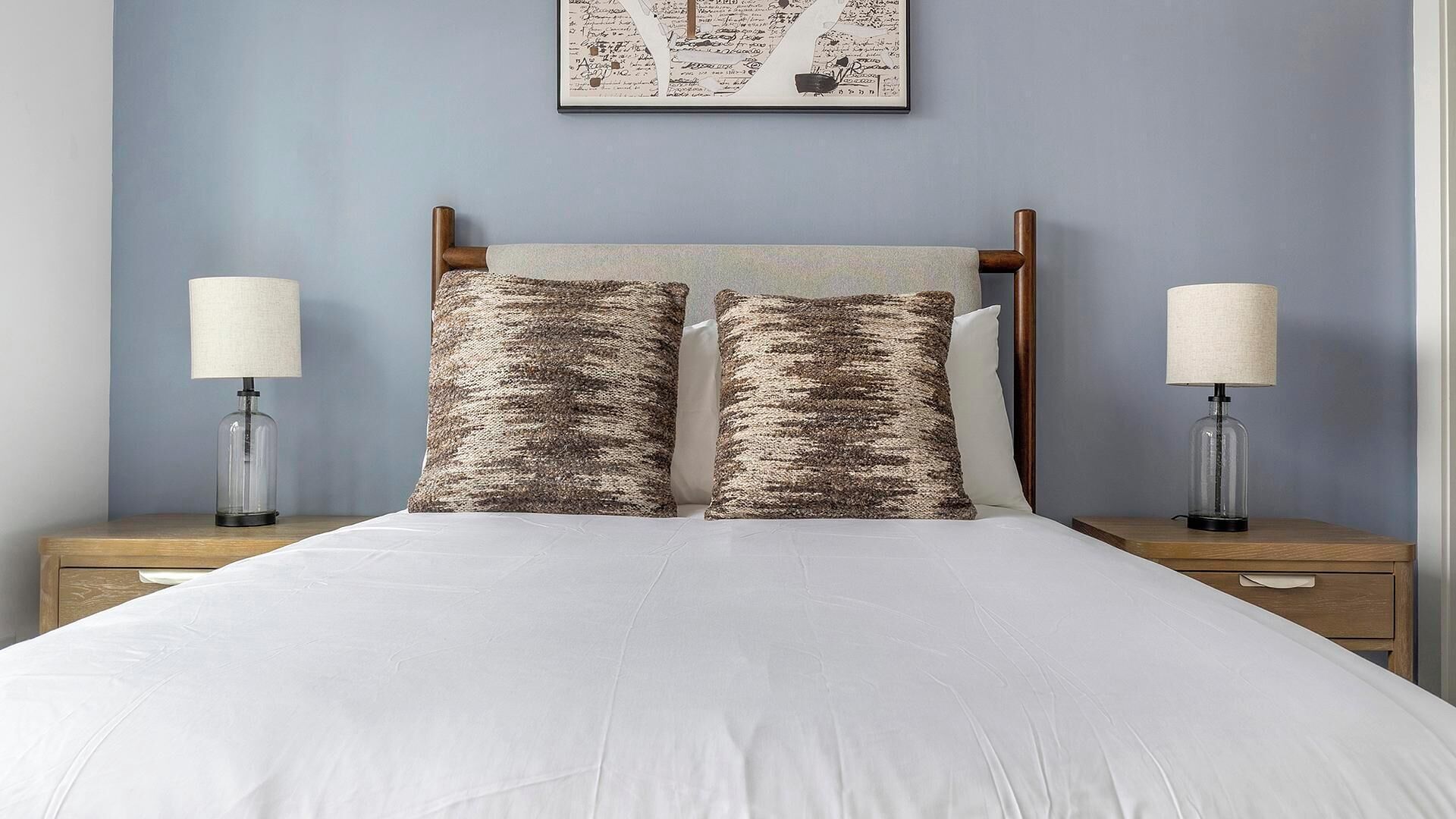 Een opgemaakt bed met witte lakens, aan weerszijden nachtkastjes met lampen en een kunstwerk erboven.