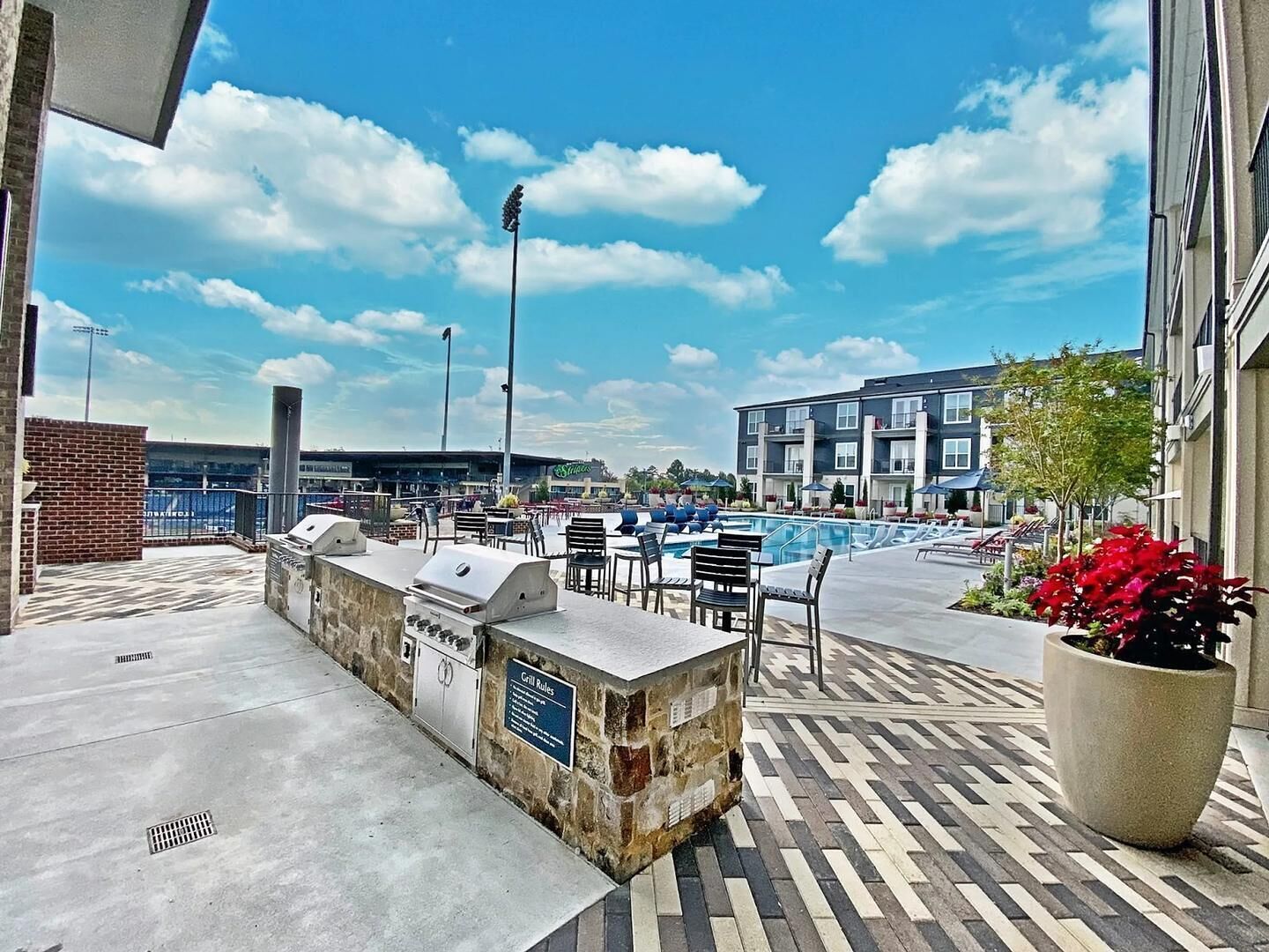 An outdoor pool area with a barbecue station, lounge chairs, and a view of a stadium in the background.