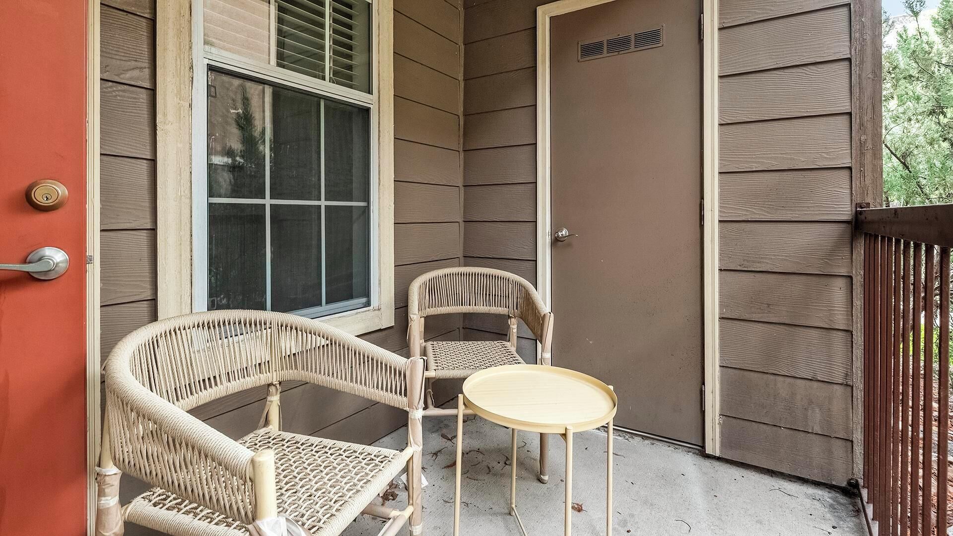 A corner patio with wicker chairs and a small round table, a sliding glass door, and a brown exterior wall.