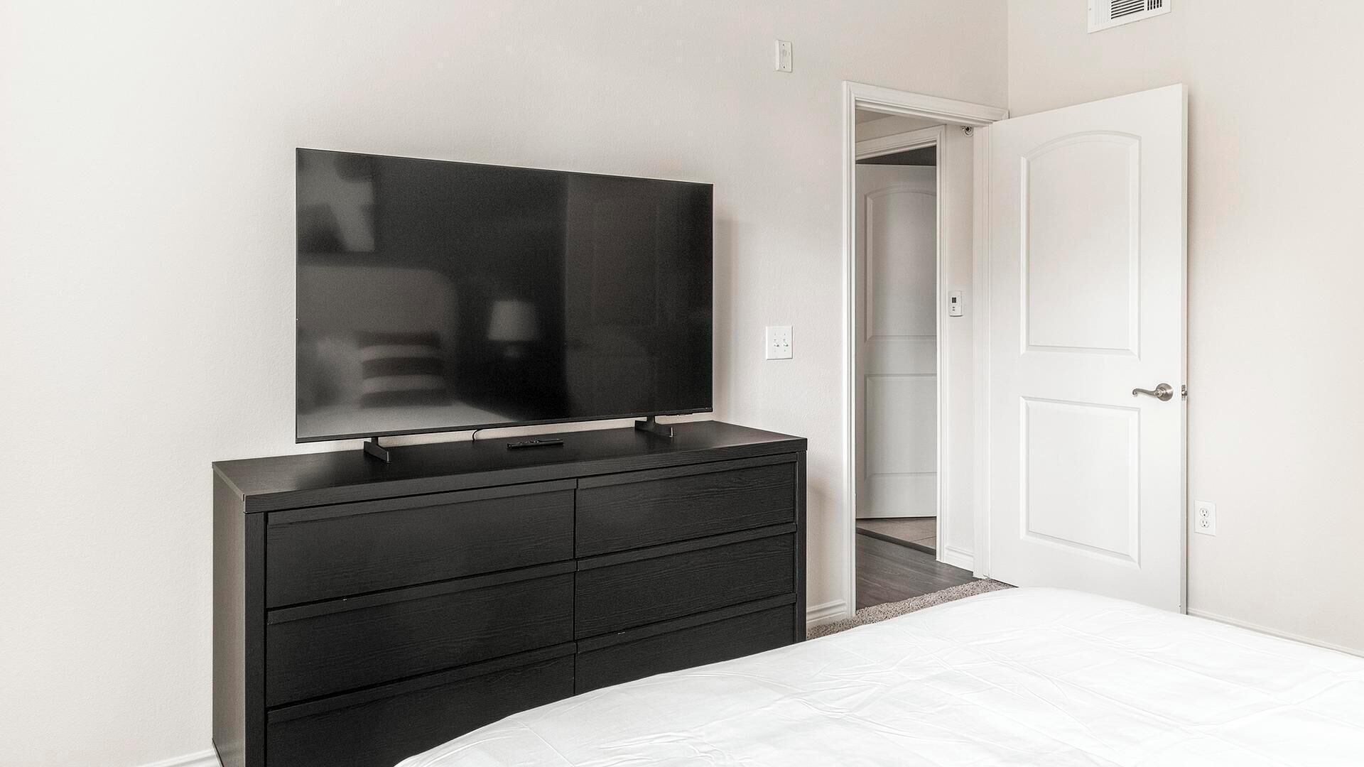 Ein Schlafzimmer mit einem großen Flachbildfernseher, der an einer dunklen Kommode befestigt ist, einer weißen Tür und einem weißen Bett.