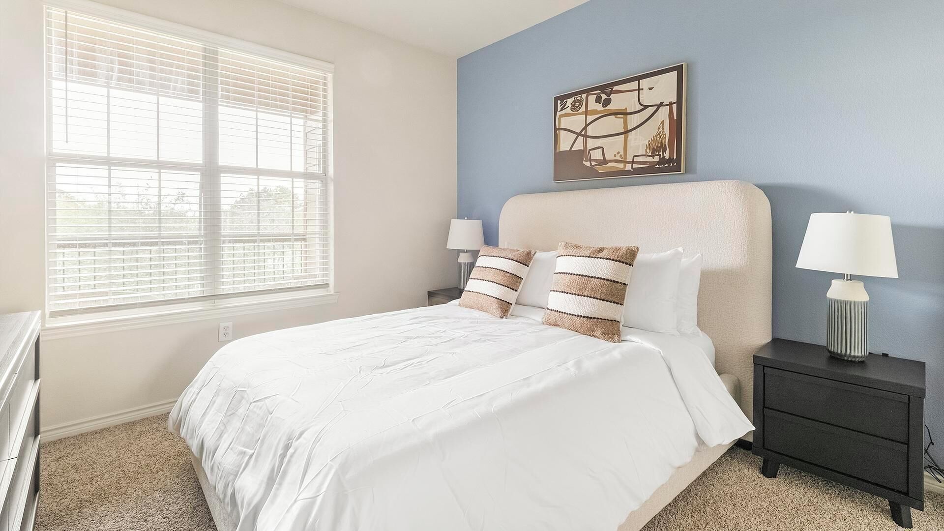 A bedroom with a bed, two bedside tables, a window with blinds, and a wall-mounted artwork.