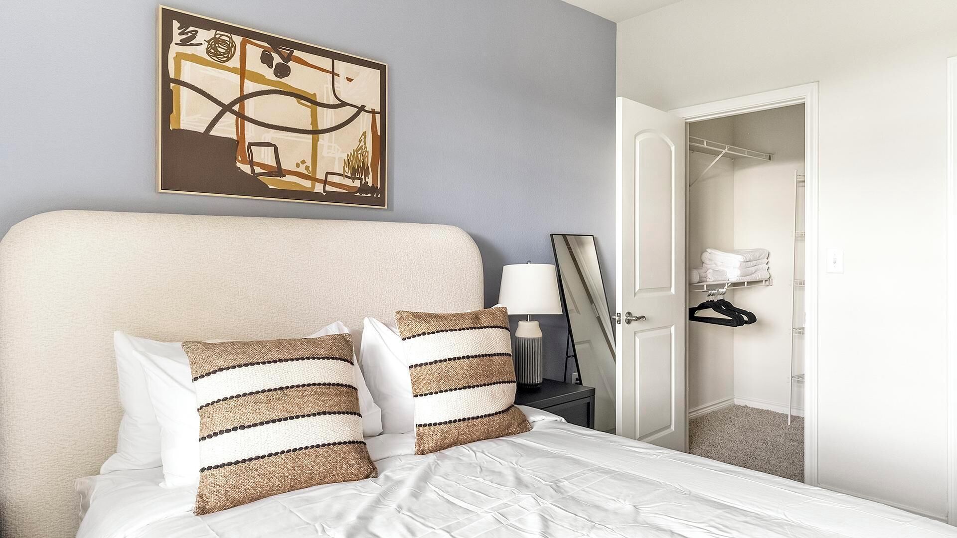 A hotel room with a bed, pillows, a nightstand, a lamp, a mirror, and a door leading to a closet with hanging clothes.
