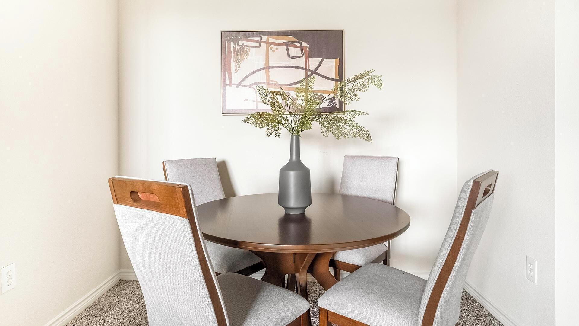 Une table de salle à manger ronde, accompagnée de quatre chaises, un vase décoré de plantes et une œuvre d’art abstraite accrochée au mur.