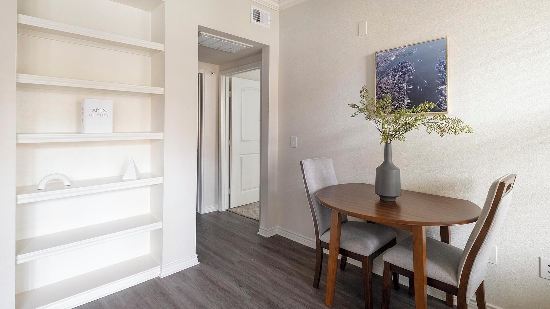 A dining area with a round table, two chairs, and a vase with flowers. There are white shelves with decorative items on the wall.