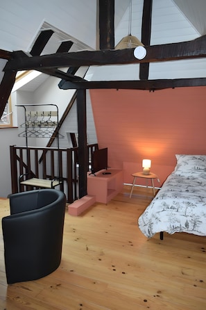 Una camera da letto mansardata con soffitto inclinato, un letto con copripiumino decorato, un tavolino con lampada, una sedia e un televisore su un mobile rosa.