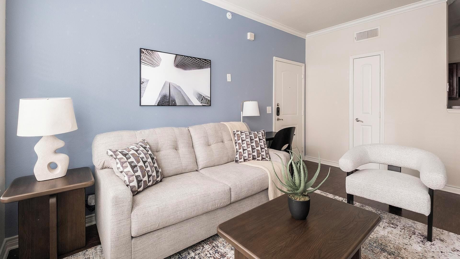 Un salon avec un canapé beige, des coussins à motifs, une table basse en bois, une plante en pot et un tableau encadré accroché au mur.