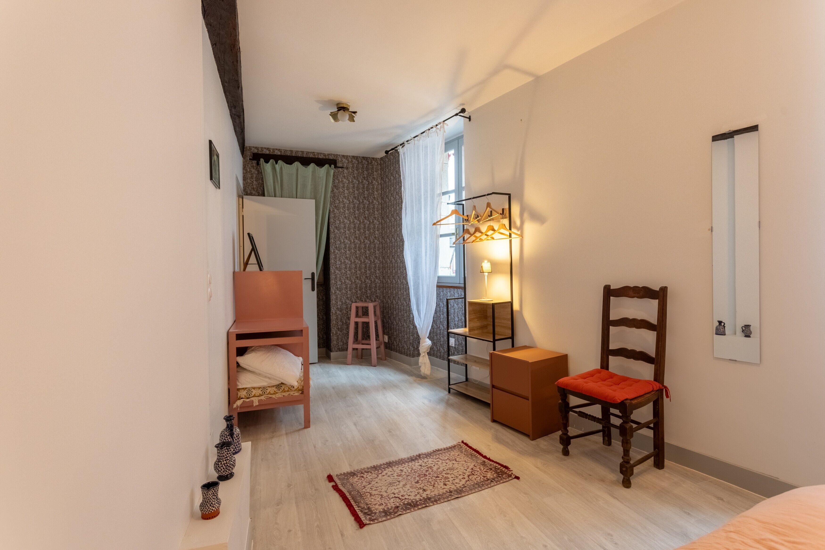 Een slaapkamer met een bed, een stoel, een bureau, een spiegel, een plank met kleding, een lamp en een raam met gordijnen.