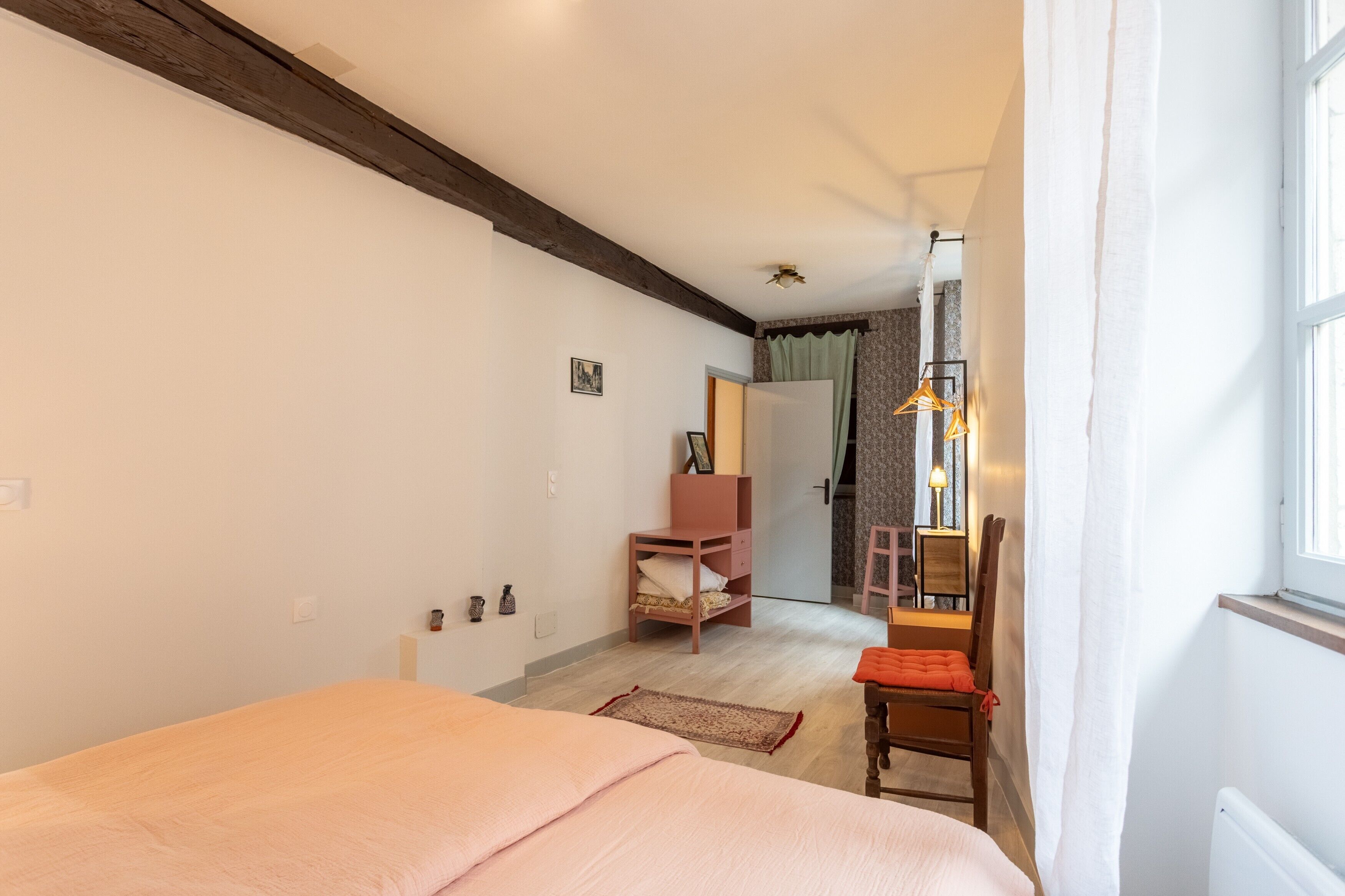 Un dormitorio con cama, una silla de madera, una mesita, un armario y una ventana con cortinas.