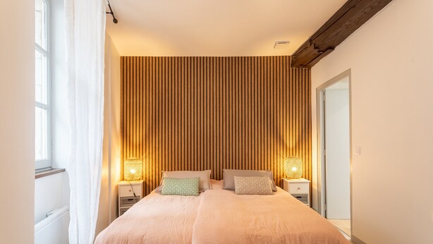 A bedroom with a bed, bedside lamps, a window, and a wooden wall panel. - Apartment 'T2 Alicia' with Wi-Fi (Salies-de-Béarn)