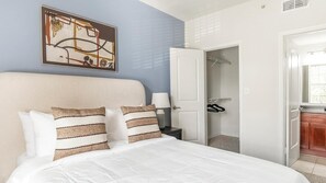 A neatly made bed with white bedding and striped pillows, a bedside table with a lamp, and a door leading to a closet with hanging clothes. - Landing | Spectacular 2BD, Gym, Clubhouse (Houston)