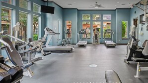 Ein Fitnessstudio mit unterschiedlichen Trainingsgeräten, großen Fenstern und einem Deckenventilator.