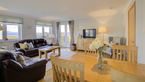 A living room with a brown leather sofa, a wooden dining table, and a television. - No 8 The Links Apartment Brora (Brora)
