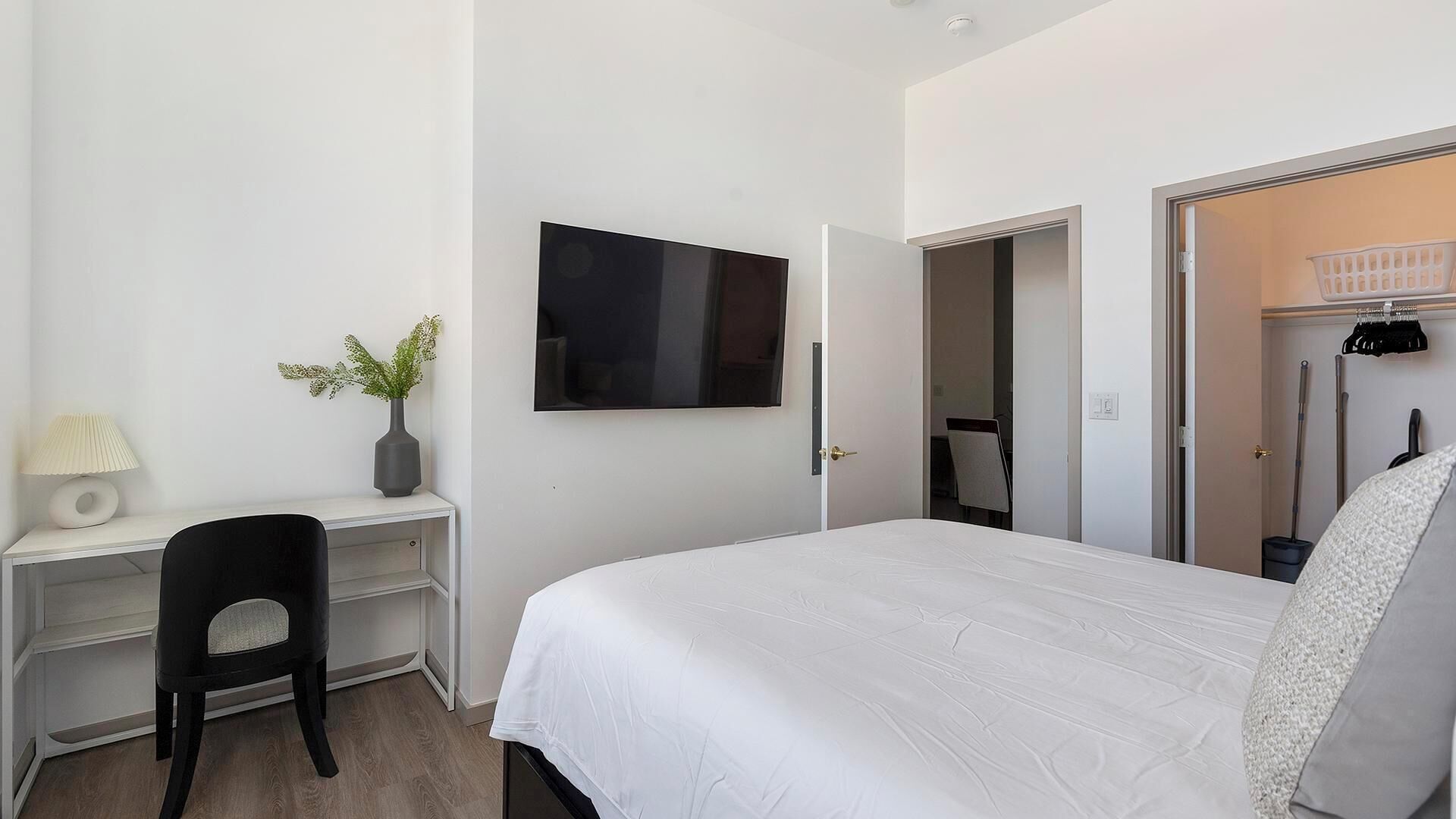 A bedroom with a bed, a desk, a chair, a TV, and a door leading to a bathroom.