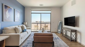 A modern living room with a sofa, armchair, TV, and a large window offering a view of the beach. - Landing | Chic 1BD, Gym, Pool (Enterprise)