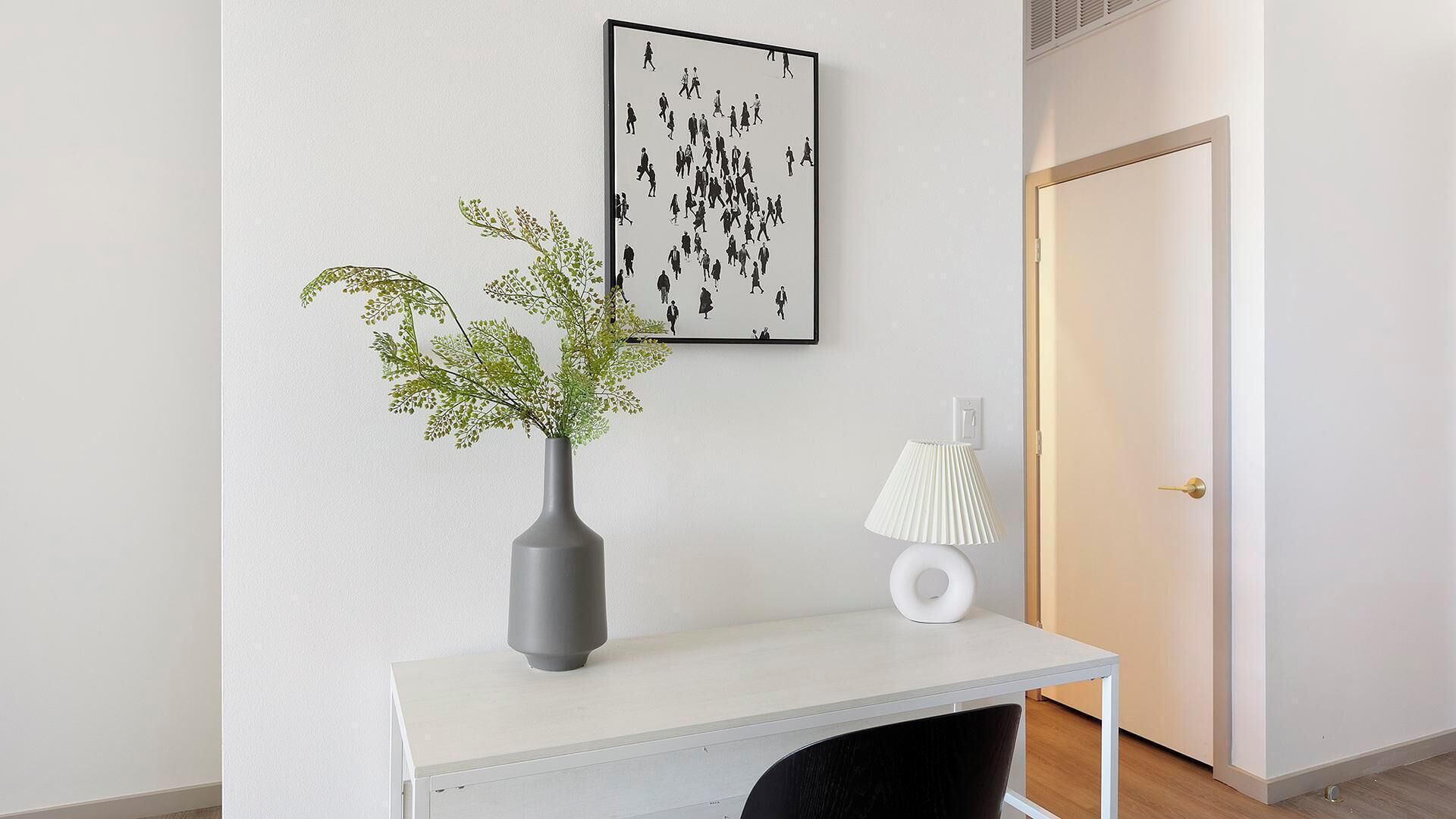 Un bureau blanc orné d’un vase gris et de quelques feuilles vertes, une œuvre d’art représentant une silhouette encadrée, une lampe blanche et une porte fermée.