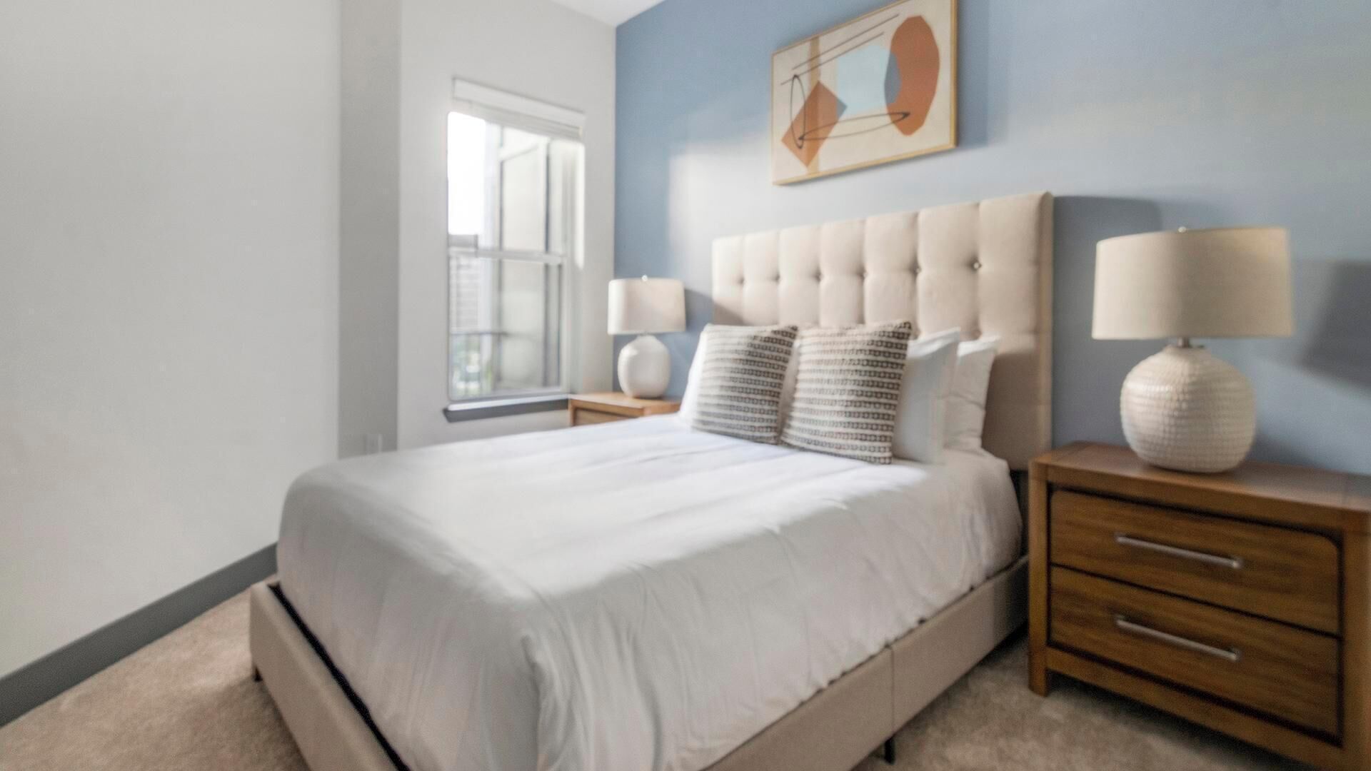 A bedroom with a large bed, a nightstand with a lamp, and a wall-mounted artwork.