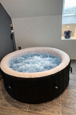 A hot tub in a room with a tiled floor, a power outlet, and a small window.