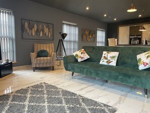 A modern living room with a striped armchair, a green sofa with floral cushions, a patterned rug, and artwork on the walls.