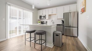 A modern kitchen with white cabinets, stainless steel appliances, a bar stool area, and a sliding glass door to the outside. - Landing | Spectacular 1BD, Gym, Courtyard (Charlotte)