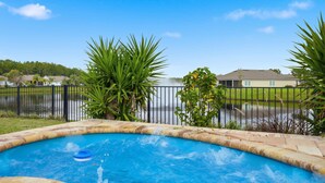 Zwembad met een jacuzzi, omringd door een hek en palmbomen, met huizen en een waterpartij op de achtergrond.