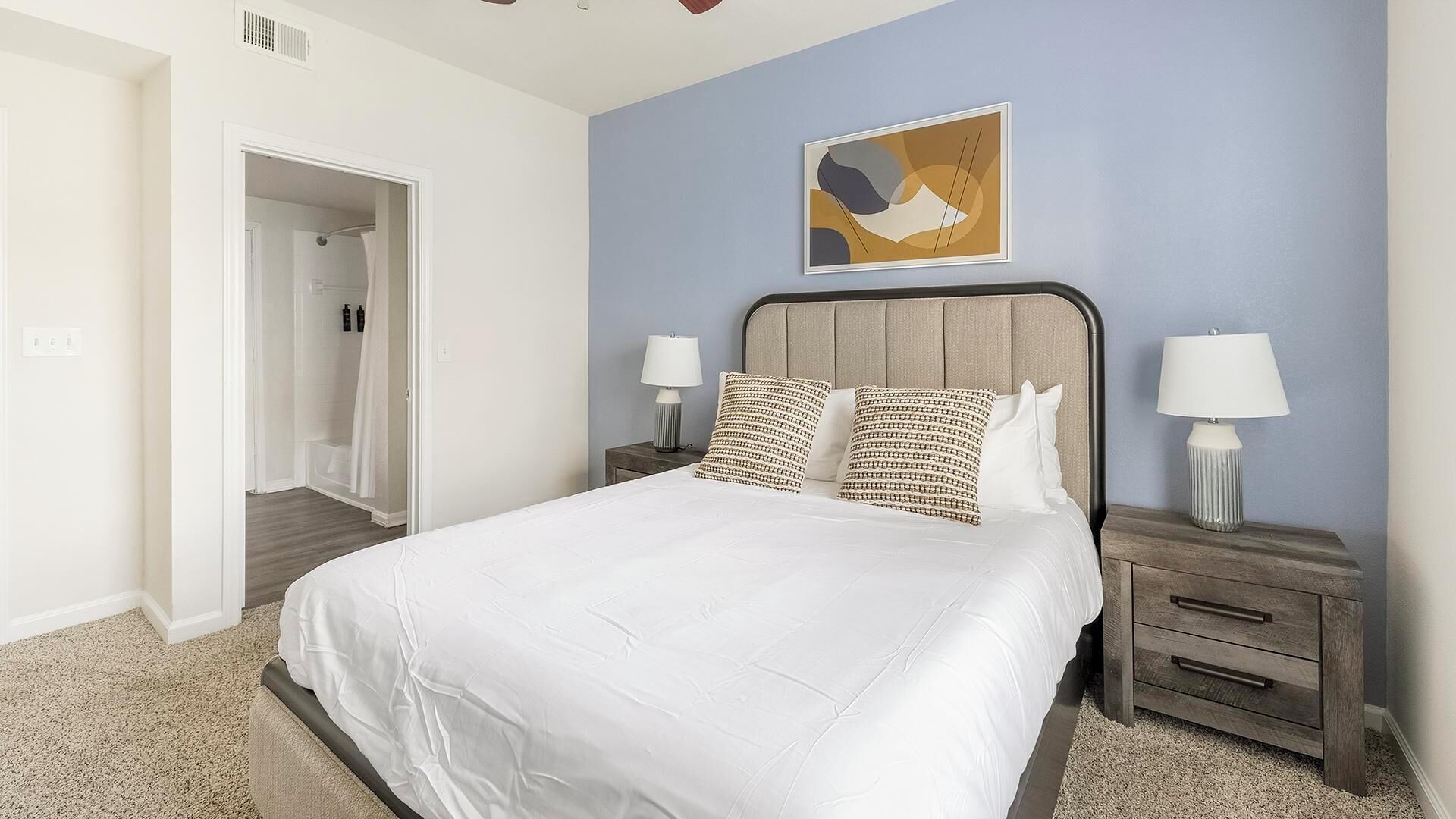 Une chambre à coucher avec un lit, deux tables de chevet avec des lampes, une porte donnant sur une salle de bain et une œuvre d’art abstraite accrochée au mur.