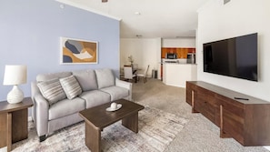 A modern living room with a sofa, coffee table, and TV. There's a kitchen area with wooden cabinets and a microwave.