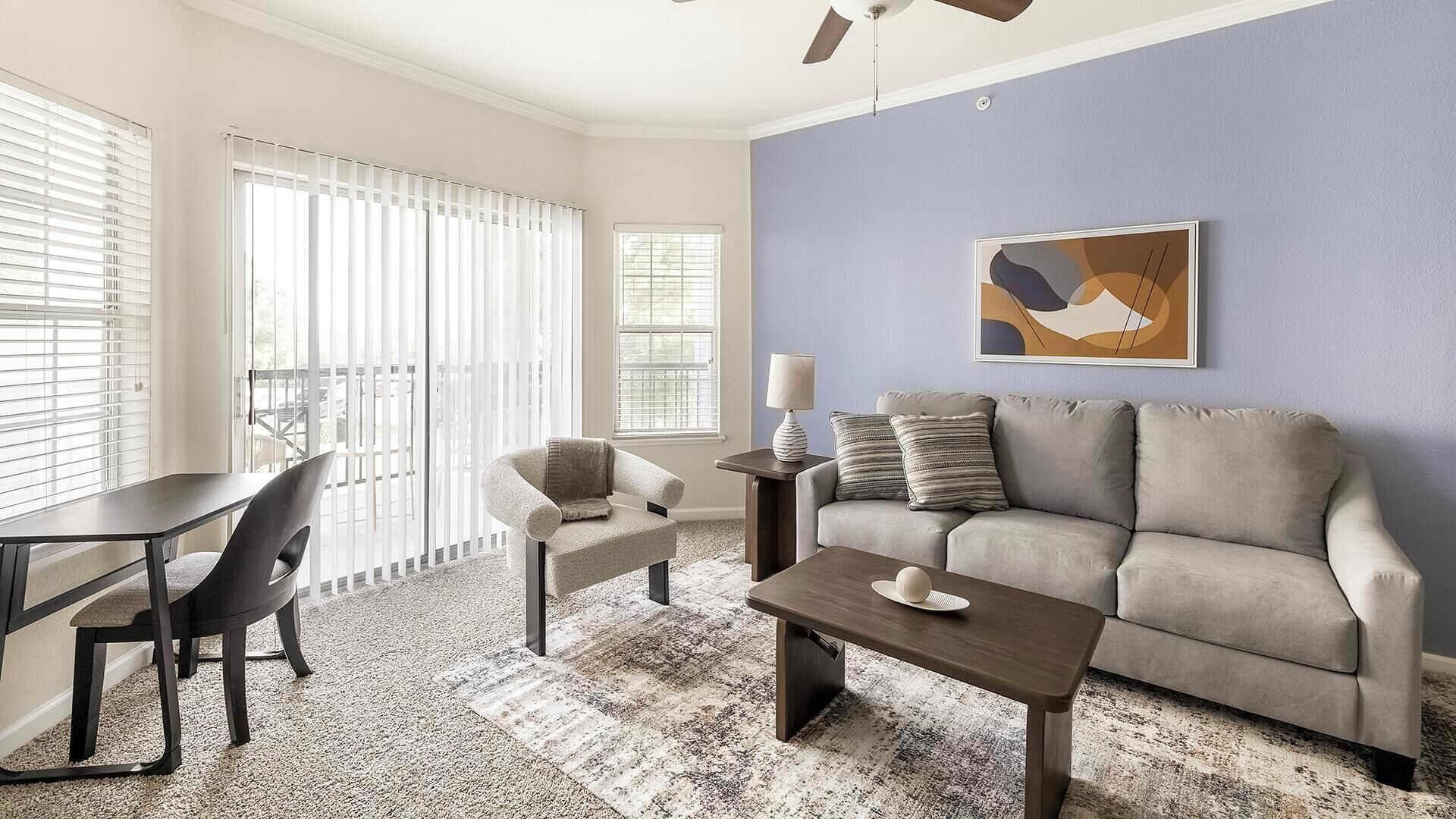 A living room with a sofa, coffee table, and a chair. There is a window with blinds, a ceiling fan, and a painting on the wall.
