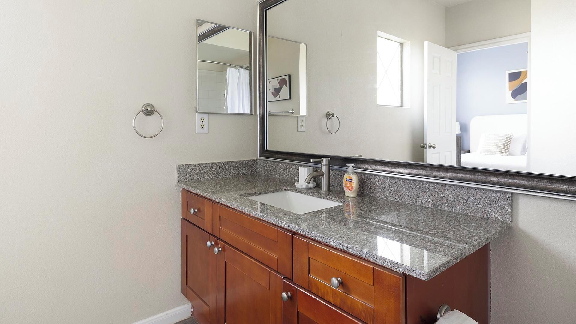 A bathroom with a granite countertop sink, wooden cabinets, a large mirror, and a window with a view of a white chair and a painting.